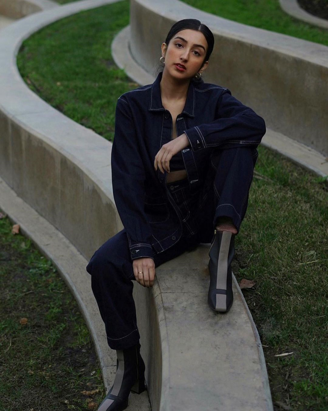 Woman in a dark denim outfit sitting on a wall made of grey stone and grass
