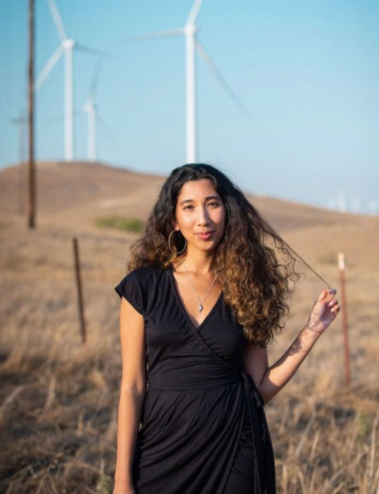 Woman wearing a black dress standing in a field, grass and wind turbines in the background.