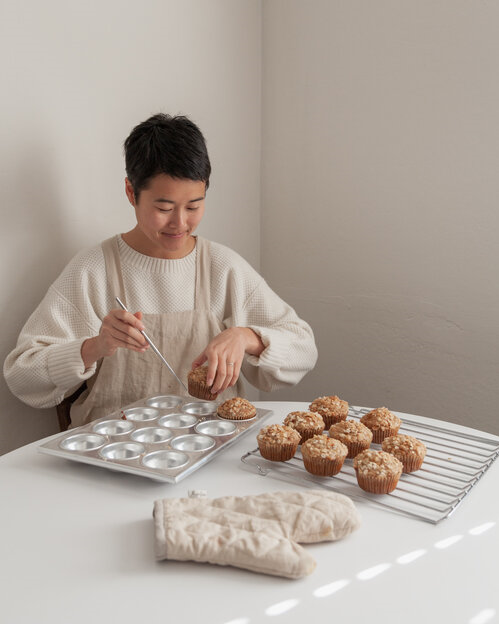 Woman in a white jumper and beige apron taking freshly baked muffins out of a baking tray
