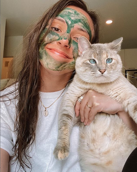 Woman wearing a green face mask, holding her grey cat up to the camera