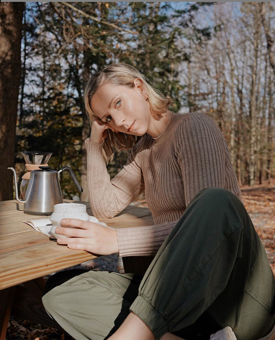 Woman sitting at a wooden table in the forest, with a kettle and a white cup in her hand.