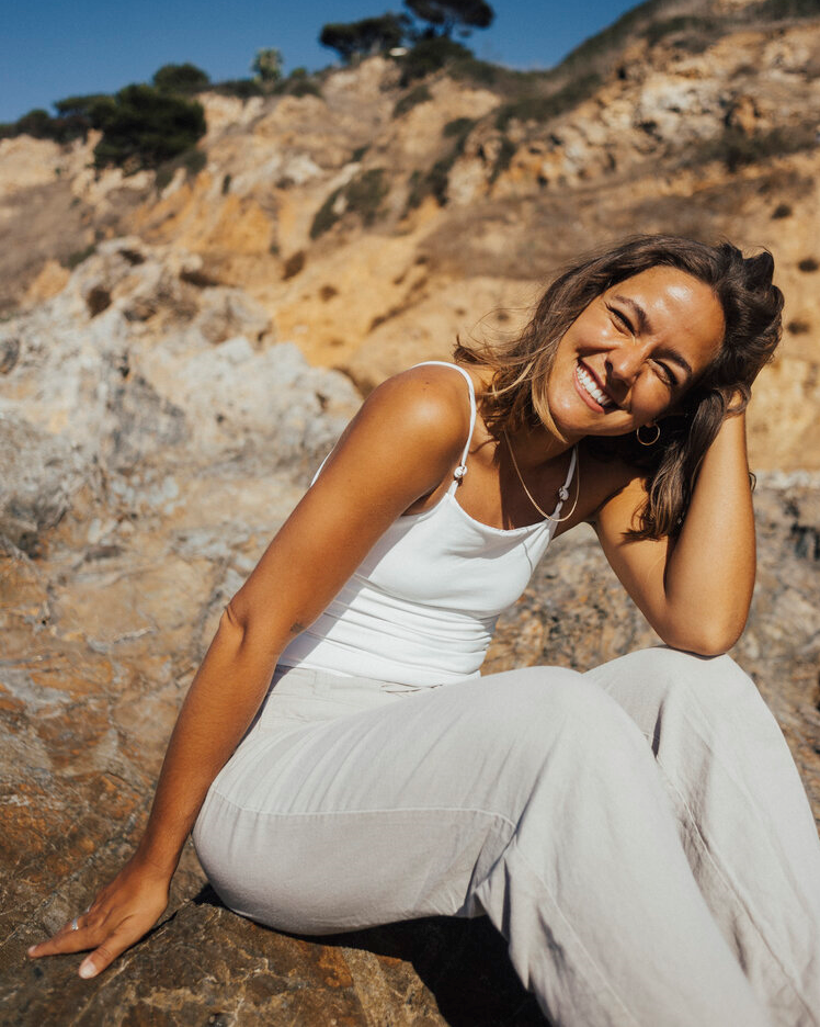 Woman smiling, in an all white outfit sitting on a rock. 