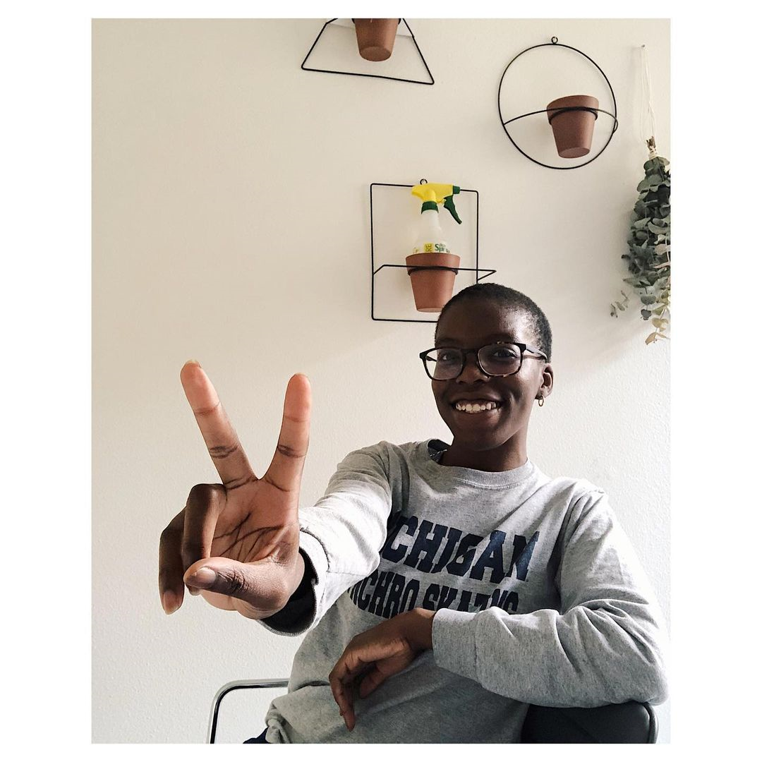 Woman smiling and giving the peace sign to a camera, behind her are plant pots on the wall