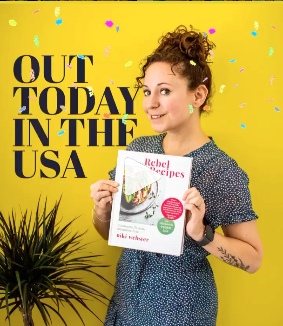A woman holding her cookbook, against a bright yellow background with the words "out today in the USA" written on the wall