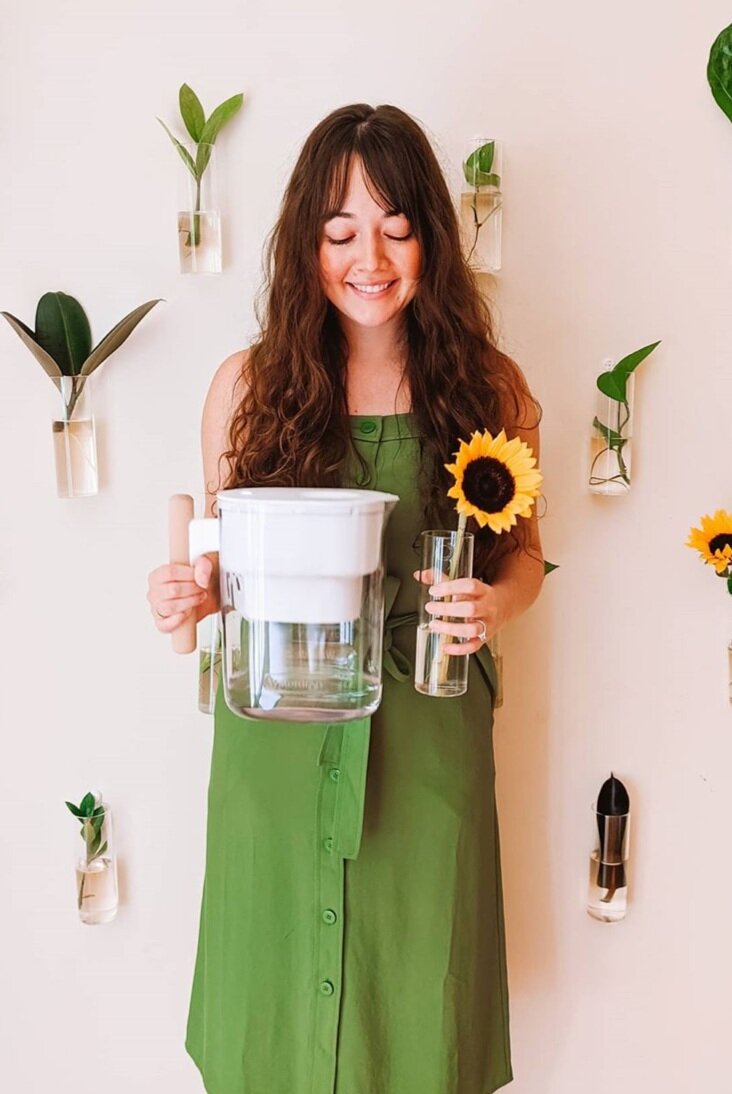 Woman in a green dress holding a jug of water in one hand and a sunflower in the other, attached to the wall behind her has different kinds of leafs in clear cups