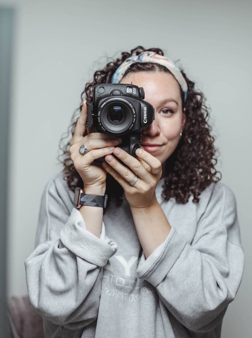 Woman in a grey jumper and colourful head band, taking a photo of someone taking her photo