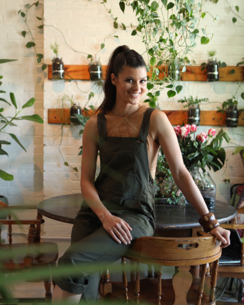 Woman in green overalls leaning on a table, Behind her are lots of plants on the floor and on the walls