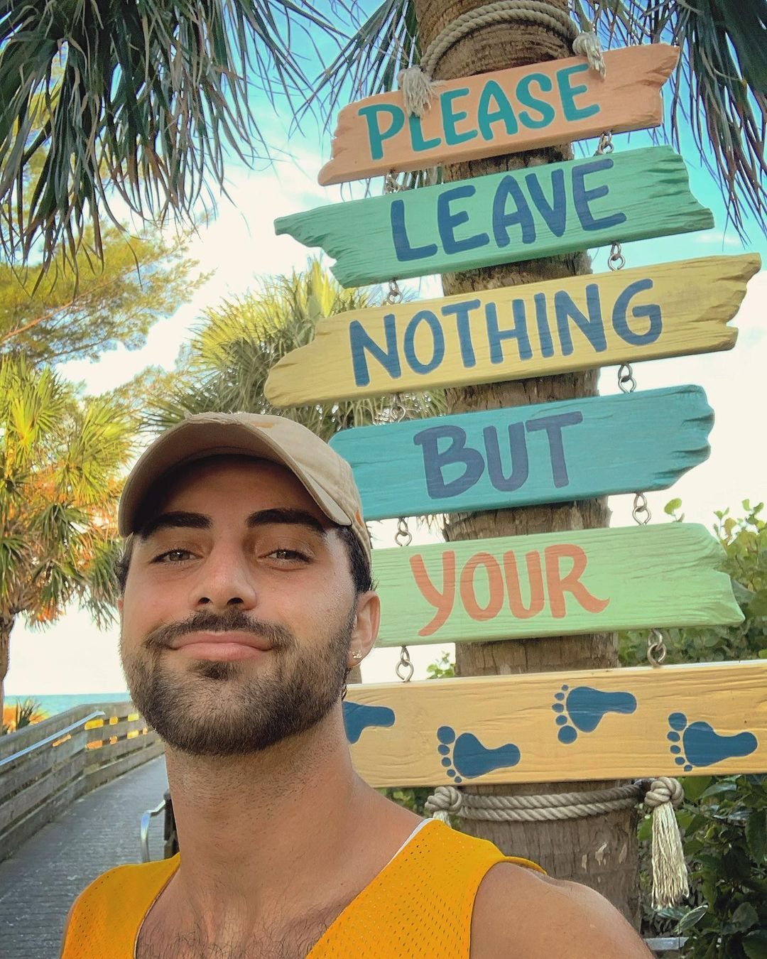Man wearing a yellow cap smiling in front of a wooden sign that reads "please leave nothing but your feet"
