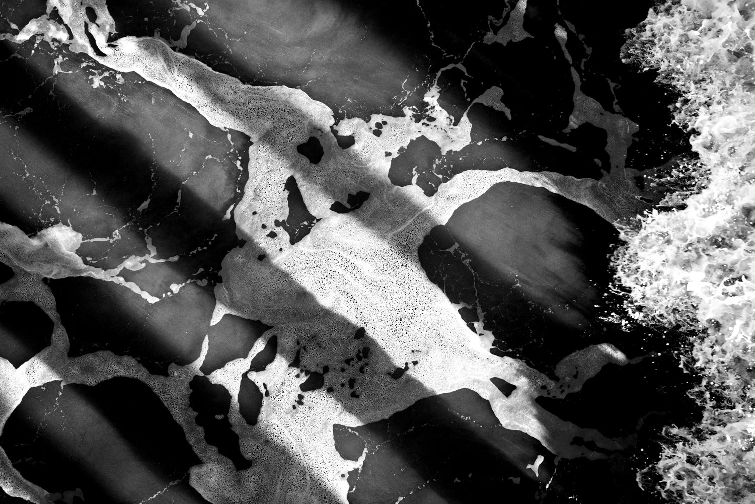 Black and white overhead view of a wave crashing onto the beach.